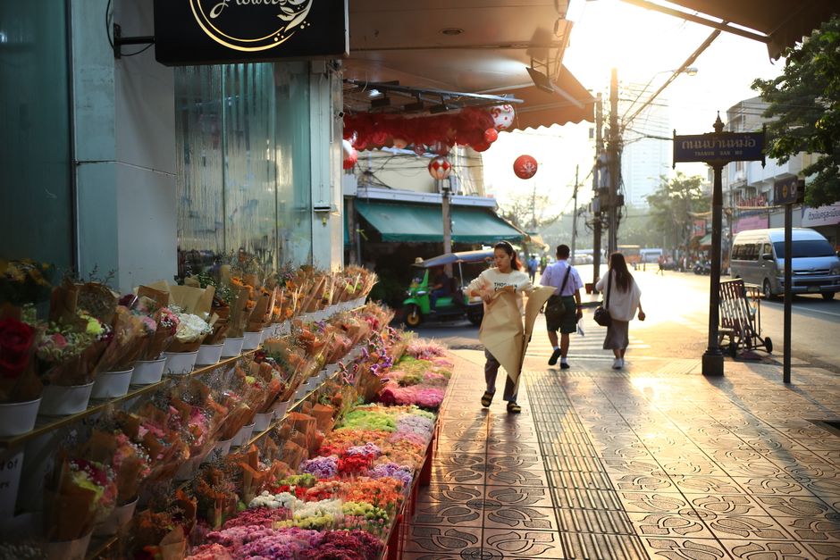A flower stall on a busy sidewalk displaying a vibrant assortment of fresh flowers, including roses, carnations, and chrysanthemums in various colors such as red, pink, yellow, and purple. The flowers