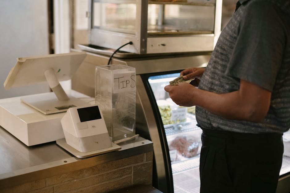 A person wearing a grey striped t-shirt stands at a cash register in a retail setting, holding a stack of banknotes ready to pay. In front of the person, the cash register and a transparent tip jar la