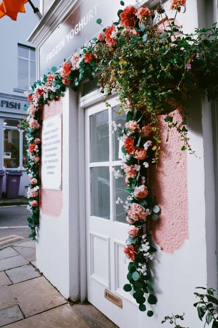 A floral arrangement of mixed pink and coral roses, complemented by smaller white and blush blooms, drapes around the white-framed window and door of a building, creating a colorful and lush floral di