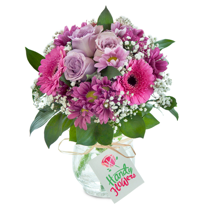 With a symphony of pink, purple, and white, this distinguished bouquet is a flourish of fresh Forest Hill blooms arranged for joyful gifting. At the core of this lush composition rest several lavender roses-each petal gently opening in soft, spiraling curves that seem to promise serenity, much like the tranquil ponds in Sydenham Wells Park. Radiant Gerbera daisies in hot fuchsia hues stand boldly around the center, their broad petals offset by dark, dramatic eyes, reminiscent of vibrant artwork in local galleries. Branches of magenta chrysanthemums intertwine, their densely packed petals and subtle green-yellow hearts adding depth and artisan texture. Wisps of white baby's breath weave through the bouquet, lightening the visual mood and creating a breathy, cloudlike finish. The folds of glossy dark green foliage beneath echo the lushness of the neighbouring woodlands. The arrangement is housed in a clear glass vase with a subtle, shimmering crackle effect, capturing the afternoon light much like windows in Devonshire Road's Victorian homes. Tied with a rustic raffia ribbon, the neck is finished with a neat white gift tag-waiting for a message to bring a Forest Hill smile to any recipient. Every element comes together in harmony, making this bouquet the perfect choice for commemorating birthdays, anniversaries, or simply brightening up a cherished home in SE23.