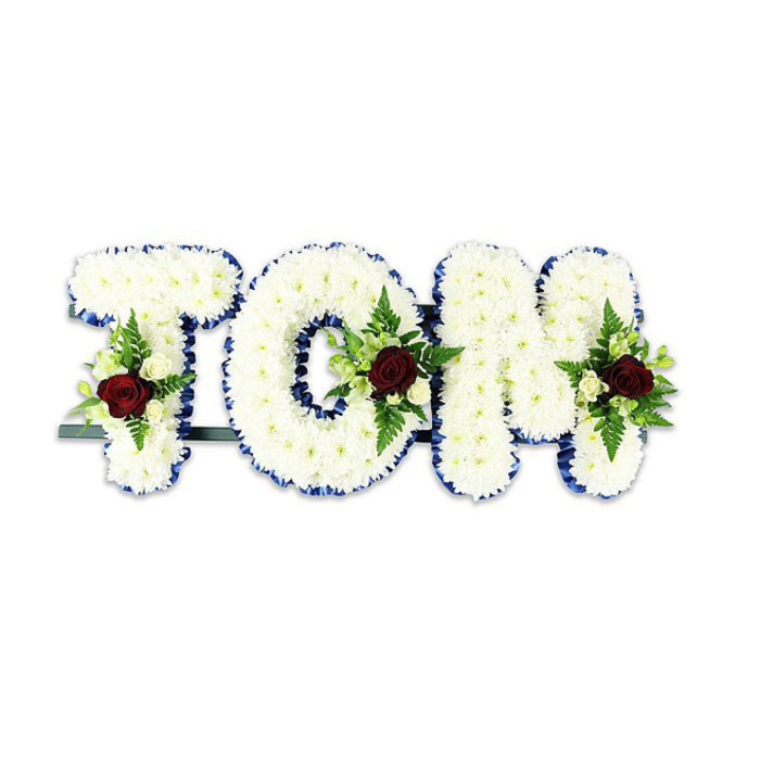 Crafted with the quiet authority of an experienced Forest Hill florist, this bespoke tribute spells TOM in three-dimensional letters entirely cloaked in small, pristine white chrysanthemums. The blooms are placed so closely together they form a pillowed surface, evoking calm and softness under the cool, soft light of a chapel or memorial hall. Each letter's silhouette is crisply defined by a royal blue ribbon that runs the full perimeter, adding a refined, almost architectural frame to the florals. Within each letter sits a composed floral spray: a single, deeply saturated red rose anchors the cluster and speaks of abiding love; surrounding it are delicate ivory spray roses and tiny white buds that offer gentle highlights. Fern fronds and assorted green foliage weave through the accents, giving natural texture and a subtle, green scent when the arrangement is approached. A dark green or black metal framework supports the letters from below, visible at the base but deliberately understated so attention remains on the flowers themselves. Placed against a stark white background, the colours read with clarity and purpose - the white chrysanthemum faces, the royal blue edge, the crimson focal roses and the fresh greens together form a composed and compassionate message. Prepared by Forest Hill Flowers for funerals, memorial services or commemorations around Dartmouth Road, Honor Oak and the Horniman area, this arrangement balances dignity, local craftsmanship and the gentle solace of a thoughtfully arranged floral name tribute.