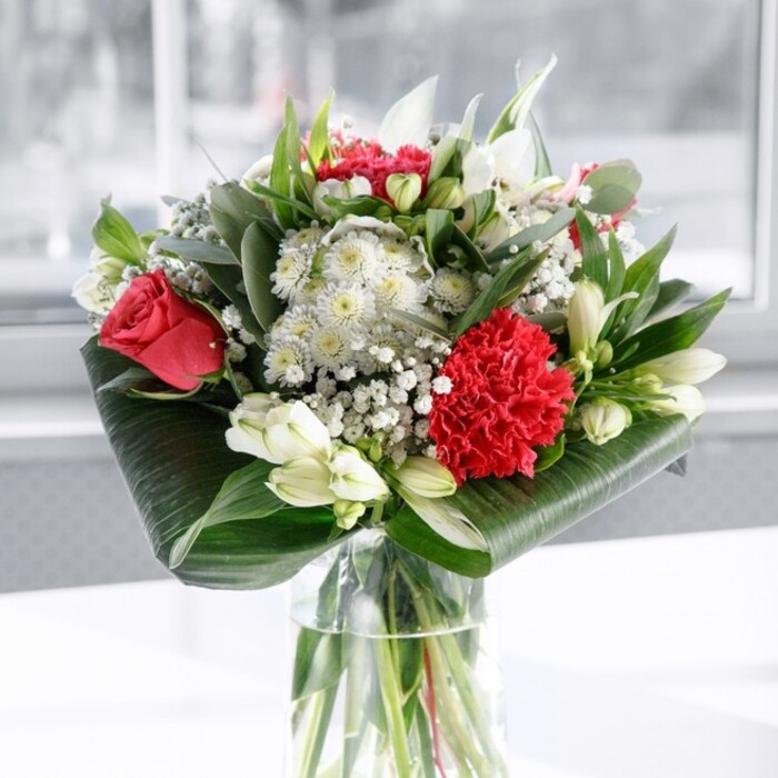 In this image, a sophisticated red and white bouquet commands attention from the centre of a clear cylindrical glass vase, its design the work of an experienced florist who understands both structure and sentiment. The vase stands on a pristine white surface against a softly blurred, pale background, reminiscent of the gentle natural light that filters through windows overlooking Forest Hill and the green slopes of nearby parks. Through the glass, you can clearly see neatly trimmed green stems submerged in fresh water, arranged in a way that feels both natural and tidy, hinting at the bouquet's longevity. Emerging from this base is a lush display dominated by deep red roses, their velvet-like petals spiralling outward, and a large, ruffled crimson carnation at the front whose intricate layers lend texture and depth. Interwoven among these rich reds are crisp white flowers: alstroemeria with fine speckles at their throats, daisy-like blooms offering a soft, open contrast, and several closed white lily buds standing gracefully upright, promising to open slowly over the coming days. Fine sprays of white gypsophila float between the larger blossoms, adding an airy, cloud-like lightness and softening the transitions between colours. Embracing the flowers is a generous collar of glossy dark green tropical leaves, carefully wrapped and curved to frame the arrangement, while additional slender, pointed leaves and eucalyptus-like foliage shoot upward and outward, creating movement and a variety of green tones. The lighting is gentle and even, picking up the sheen on the leaves and the delicate textures of each petal. The overall impression is one of refined romance and thoughtful composition, ideal as a centrepiece for a family gathering in a Forest Hill home, a warm welcome in a local office, or a heartfelt gift that conveys admiration, gratitude, or love.