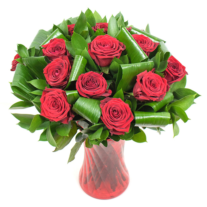 A sumptuous bouquet of approximately a dozen deep crimson roses is shown here, each flower in full, perfect bloom with velvety, tightly spiralled petals that suggest freshness and classic romance. The rose heads are closely arranged into a lush, rounded mass, their rich, wine-red tones intensified by soft, even lighting that reveals the delicate layering and natural sheen of each petal. Interwoven among the roses is a generous volume of dark green foliage: smaller, glossy leaves forming a dense verdant backdrop and larger, more lustrous leaves folded into sculptural tubes and sweeping curls. These folded elements-often Aspidistra or similar broad-leaf material-are manipulated into elegant loops that cradle the blooms and introduce an architectural, contemporary sensibility to a timeless subject. The bouquet is housed in a tall, translucent ruby-red glass vase with a subtle twisted or ribbed surface that captures vertical bands of reflected light; its narrow waist and flared top give the design a poised, statuesque quality. Set against a pristine white background, the reds and greens read with heightened clarity and emotional warmth. This arrangement suggests intimate occasions-anniversaries, Valentine's Day, or a refined romantic gift-and the scent of fresh roses seems almost perceptible. Picture this design thoughtfully delivered to a Forest Hill flat overlooking Horniman Museum & Gardens, where the intense red would sing against a grey London sky and warm a room with classic charm.