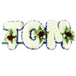 The photograph shows a traditional funeral name tribute spelling out TOM in broad, three-dimensional letters constructed from pristine white chrysanthemums. The blossoms are packed closely together to form a thick, uniform layer of petals, giving each letter a cushiony, tactile feel that is both gentle and reassuring, echoing similar memorial pieces often delivered to Forest Hill and surrounding areas on the morning of a service. Around the outer edge, a royal blue satin ribbon runs in a clean, pleated line, tracing the neat contours of the T, the rounded O, and the strong M, while its subtle sheen adds depth and formality against the simple background. Breaking the sweep of white in three places are carefully positioned floral accents: on the top-left of the T, along the left side of the O, and at the top-right of the M. Each cluster contains velvety red roses, their petals rich and full, joined by smaller white or cream spray roses and graceful, narrow white blooms reminiscent of freesia. These are supported by pockets of greenery-airy fern fronds, smooth leaves, and slightly darker foliage-creating contrast and a natural sense of movement as they rest on the cushion of chrysanthemums. The letters sit firmly on a slim, dark frame that holds them in a neat horizontal row, suitable for placement by a coffin, on a hearse, or at the head of a memorial display. This tribute combines purity, respect, and emotional warmth, making Tom's name the quiet, dignified focus for friends and family gathering to remember him around Forest Hill.