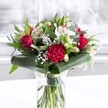 A dramatic red and white bouquet stands proudly in a clear cylindrical glass vase, set on a clean white surface with a softly blurred, light-filled background that suggests a bright Forest Hill living room or studio near the station. Inside the vase, you can see the fresh green stems neatly trimmed and fanned out below the waterline, a sign of careful conditioning and same-day preparation. The arrangement is built around velvety deep red roses, their petals beginning to unfurl in luxurious spirals, mingling with a large, ruffled crimson carnation at the front whose frilled edges add a rich, tactile texture. Threaded between the red blooms are elegant white alstroemeria, their narrow petals marked with fine speckles at the throat, along with white chrysanthemum or daisy-style flowers and several closed white lily buds, their smooth, tapered forms hinting at fragrant blossoms still to come. Clusters of delicate white gypsophila, or baby's breath, drift through the arrangement like a soft mist, lightening the palette and enhancing the sense of fullness. Around the base, broad, glossy dark green tropical leaves have been artfully curled to form a structured collar, while additional slender leaves and eucalyptus-like foliage weave upwards, providing varied greens and subtle movement. The light falls evenly across the bouquet, catching the shine on each leaf and the softness of every petal, creating an inviting, celebratory mood. The overall impression is of classic romance with a modern edge, ideal for an anniversary dinner in a Forest Hill apartment, a birthday surprise delivered along Dartmouth Road, or simply to bring a warm, heartfelt presence into an office or home.