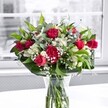 A lush, romantic bouquet of deep crimson red roses, matching red carnations, and crisp white alstroemeria stands proudly in a classic clear glass vase on a pure white tabletop, bathed in soft daylight from a nearby window overlooking Forest Hill. The roses, some fully open with velvety petals spiralling outwards and others still in tight, promising buds, create a sense of unfolding love and celebration. Nestled among them, plump ruffled carnations in the same rich red tone add fullness and a slightly old-fashioned charm, their many-layered petals catching the light in gentle shadows. Between the bold reds, clusters of white alstroemeria-some buds, some partially unfurled-show their slender, trumpet-shaped blooms tipped with a whisper of green, suggesting freshness and quiet elegance. Sprays of tiny white gypsophila, or baby's breath, form a misty halo throughout the arrangement, softening the contrast and giving the bouquet an airy, cloudlike texture. Surrounding the flowers, glossy deep green leaves and slimmer, lighter greenery arch and weave through the design, framing the bouquet with a natural, sculpted collar that makes the arrangement feel both polished and generous. Through the clear glass, the neatly cut stems are visible, fanning out in clean water that glints against the white surface beneath. The background, gently blurred and almost monochrome, might be a calm Forest Hill street scene glimpsed through the window, its muted tones allowing the red, white, and green flowers to stand out with striking clarity. The overall mood is one of thoughtful romance and gratitude, the sort of design a local florist might suggest for an anniversary, Mother's Day surprise, or a heartfelt thank-you delivered to homes around the SE23 hills.