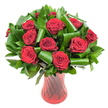 A luxurious, rounded bouquet of roughly a dozen deep crimson roses fills the frame, each bloom open to reveal tightly spiralled, velvety petals in rich wine-red tones. The rose heads sit closely clustered, their plush texture and faint classic rose scent almost tangible beneath the even, soft lighting that brings out subtle highlights along each petal edge. Surrounding and supporting the blooms is abundant, glossy dark green foliage-smaller, dense leaves that form a verdant backdrop and larger, emerald-green leaves that have been expertly folded and rolled into tubular, architectural shapes. These sculpted leaves bend and curl in elegant loops, framing individual roses and lending a contemporary, structured silhouette to an otherwise traditionally romantic composition. The whole arrangement is presented in a tall, translucent ruby-red glass vase with a delicate ribbed or twisted pattern that catches and reflects light in vertical streaks, the vase's slender waist and flared lip visible against a crisp white background. That pure white field isolates the colours so the reds read deeper and the greens read fresher, allowing the bouquet to read as both a heartfelt gift and a refined centrepiece. The overall mood is one of classic passion balanced with modern design-a bouquet that would suit an anniversary, Valentine's gift, or an intimate celebration. As a local touch, imagine this arrangement displayed in a Forest Hill flat near the Horniman Gardens, where the deep crimson would contrast beautifully with early evening light through the windows.