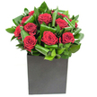 In this elegantly modern photograph, a compact cluster of rich crimson roses commands the center with velvety, tightly spiralled petals that suggest both intimacy and theatre. Each bloom is planted close to its neighbour, creating a dense heart of red that glows against the cool clarity of surrounding foliage. The leaves are broad, glossy and expertly curled into overlapping loops, their emerald sheen catching the steady studio light and creating a rhythmic, almost architectural pattern around the roses. The arrangement sits in a matte black cube - a low, square vessel whose flat planes and grounded silhouette lend contemporary poise, balancing the lushness above with a minimal, urban attitude. Light is clean and even, skimming the tops of petals and throwing delicate highlights along the waxy leaf edges so that texture reads as clearly as colour. Run a fingertip near the display in your imagination and you'd sense the contrast: the soft, pillowy give of rose petals against the firmer, almost tropical bite of the greenery. There is a faint, fresh fragrance - the green, leafy notes of newly cut stems mingling with a classic, sweet rose perfume - evocative of a quiet morning market trip rather than cloying perfume. This bouquet reads as an intentional statement piece, perfect for an anniversary, a romantic milestone, or to bring considered warmth to a contemporary living room or office. The craftsmanship is precise yet soulful, a modern love letter that feels at home on a Forest Hill mantelpiece or as a thoughtful gift from neighbours who know how to marry bold colour with composed structure.